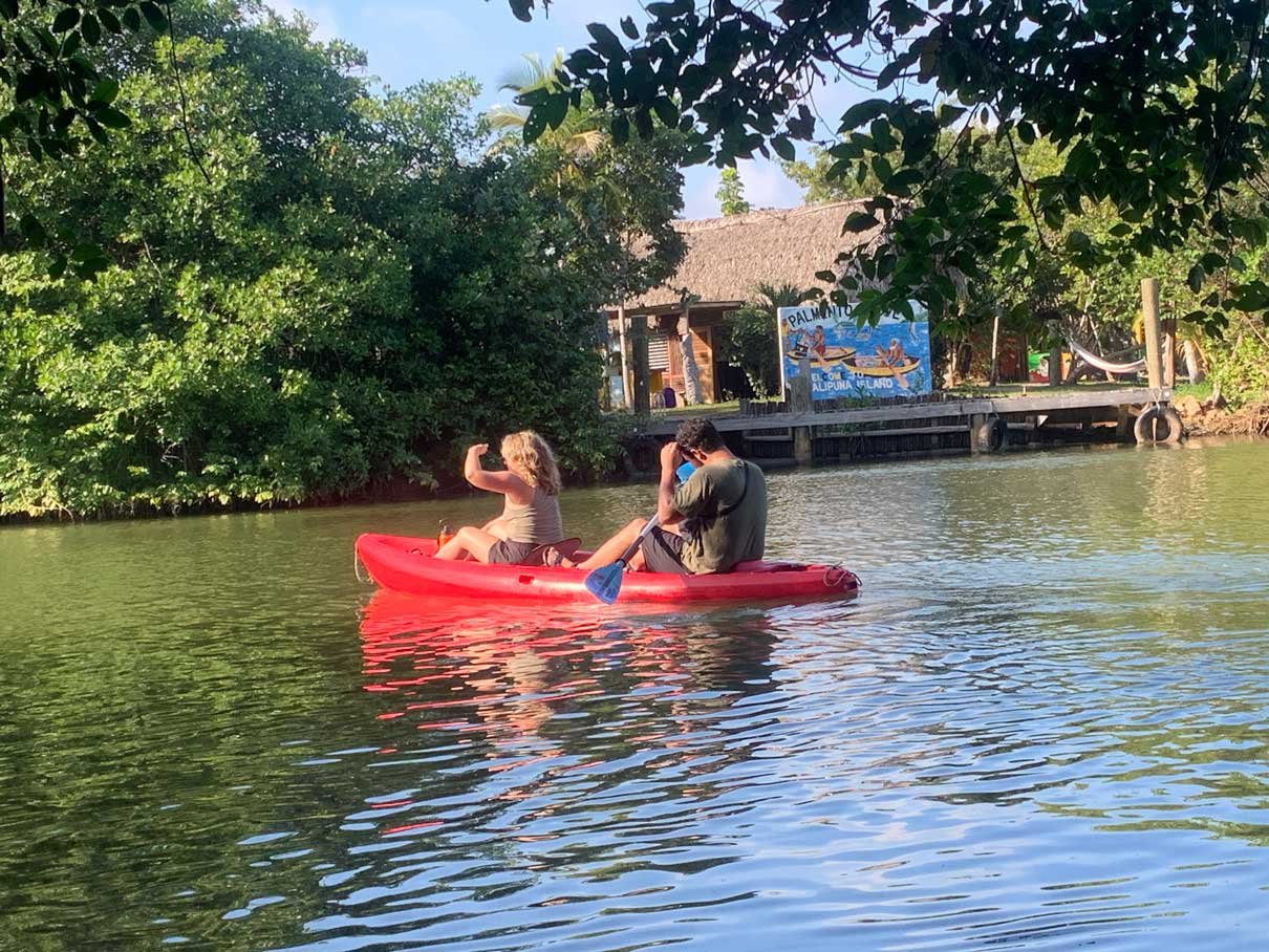 Two guests in a red kayak on the lagoon with the Palmento Grove landing behind them, Hopkins, Belize
